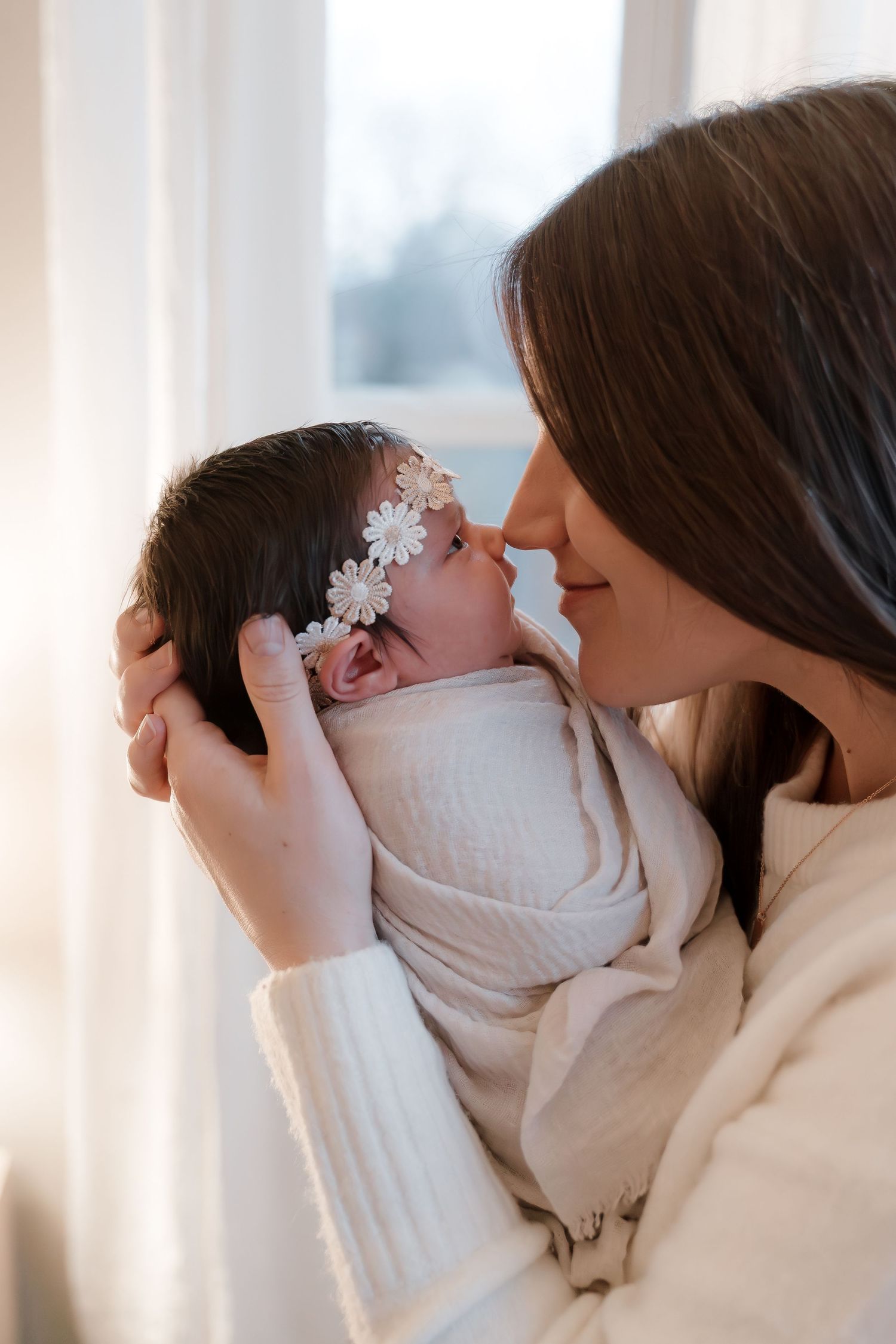 A mother tenderly holds her swaddled newborn baby girl wearing a floral headband near a bright window.