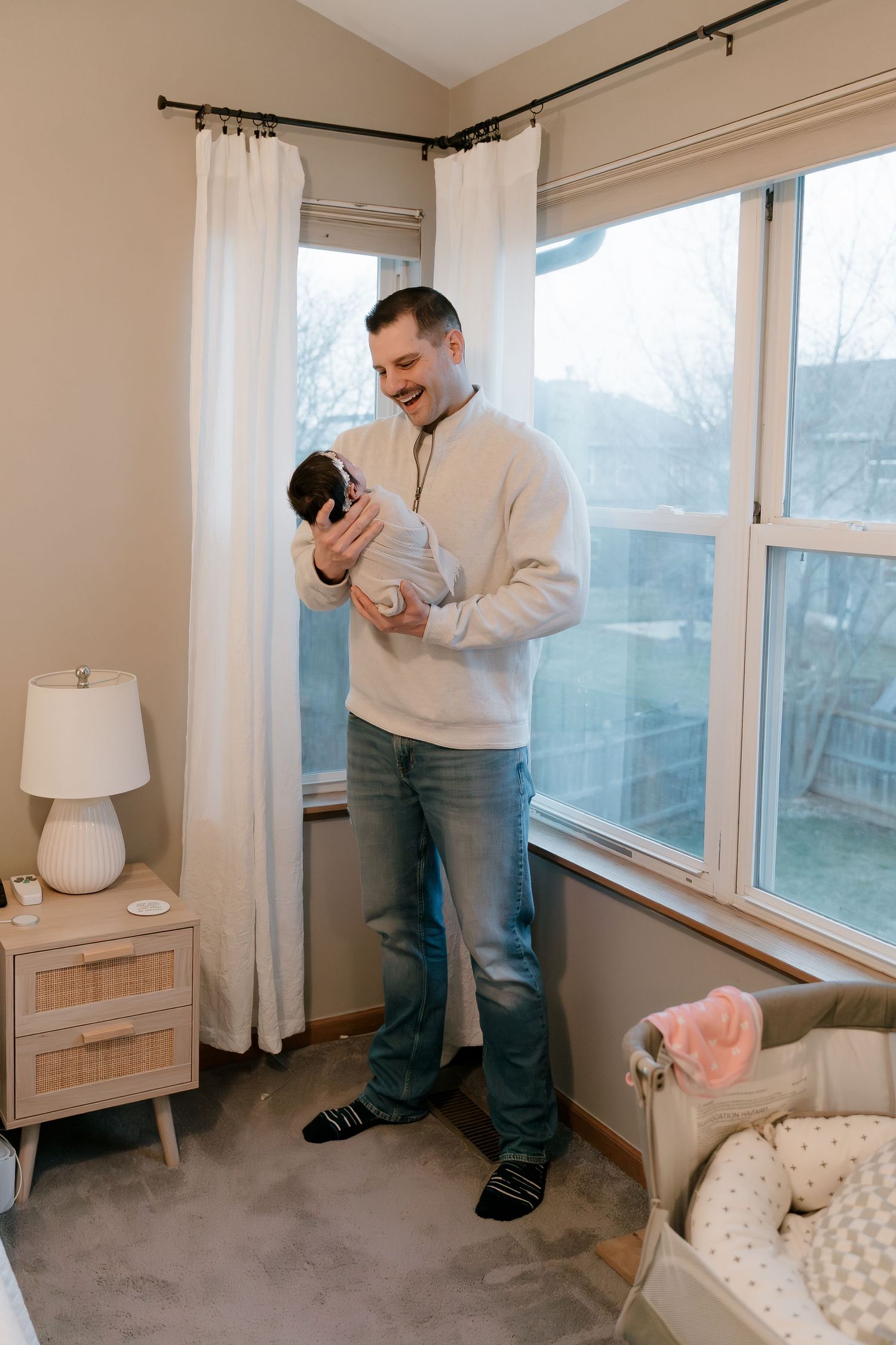 A smiling father stands by a bright window holding his newborn baby in a cozy nursery room.