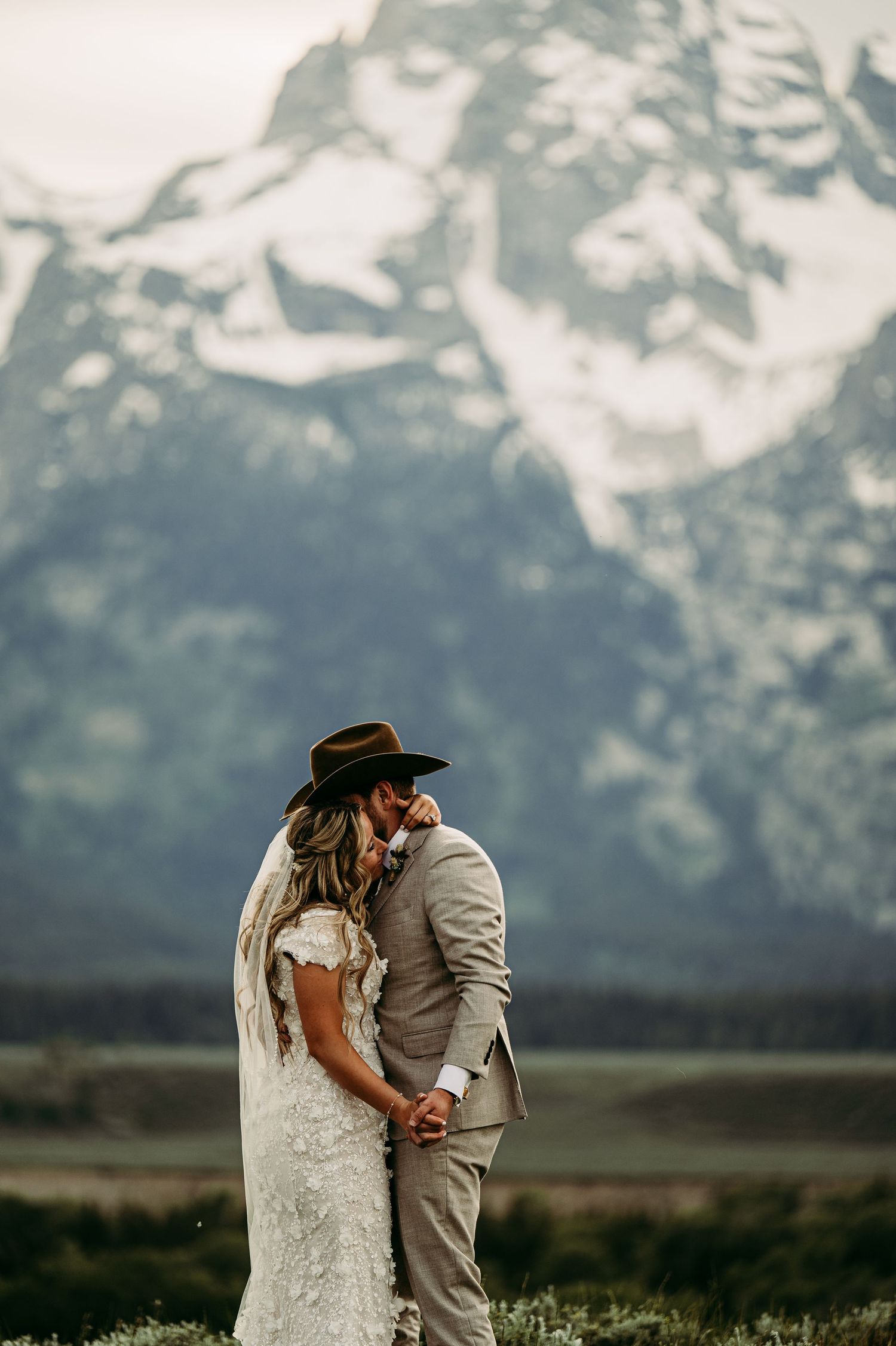 A woman with manicured nails and an engagement ring embraces a man from behind against a mountain backdrop.