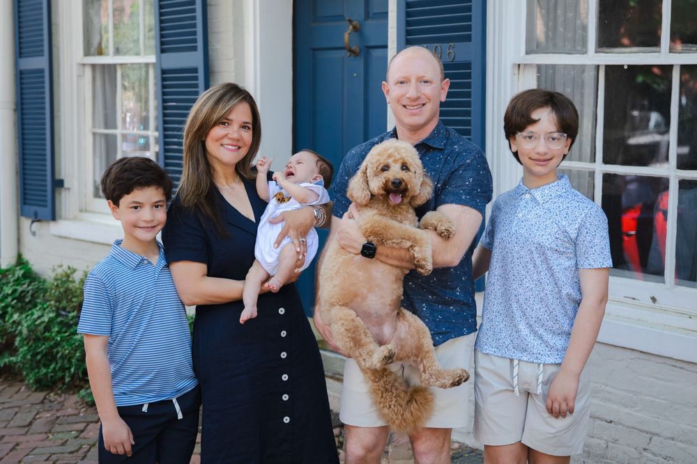 Happy family of five posing outdoors with a golden doodle dog, including two boys, parents, and a newborn baby.