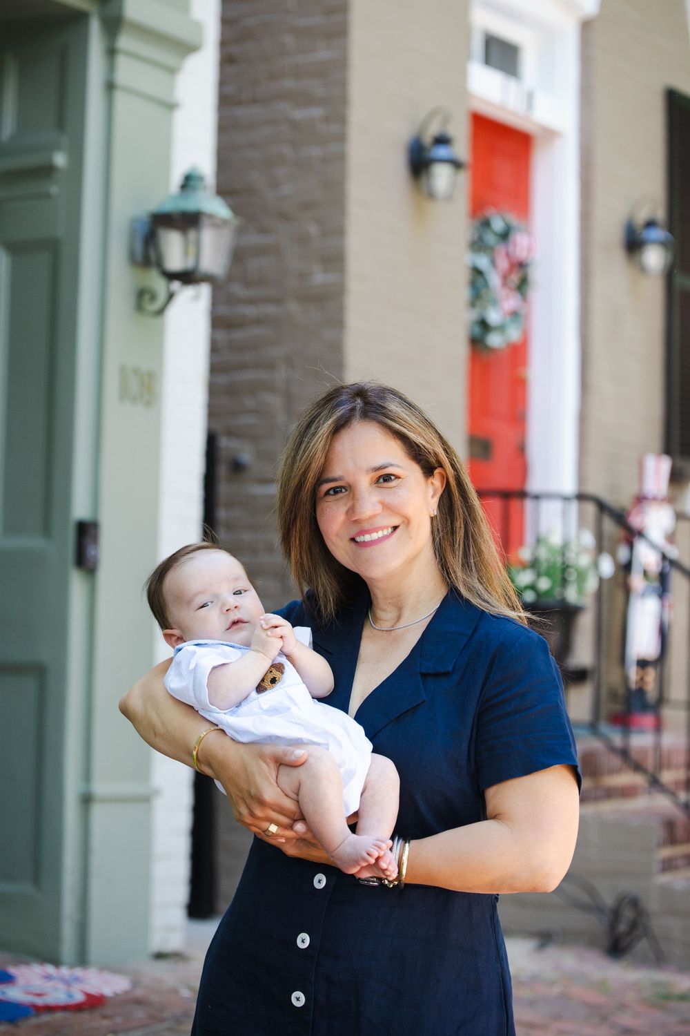 Woman in navy dress smiling while holding a newborn baby outside a brick rowhouse with red door.