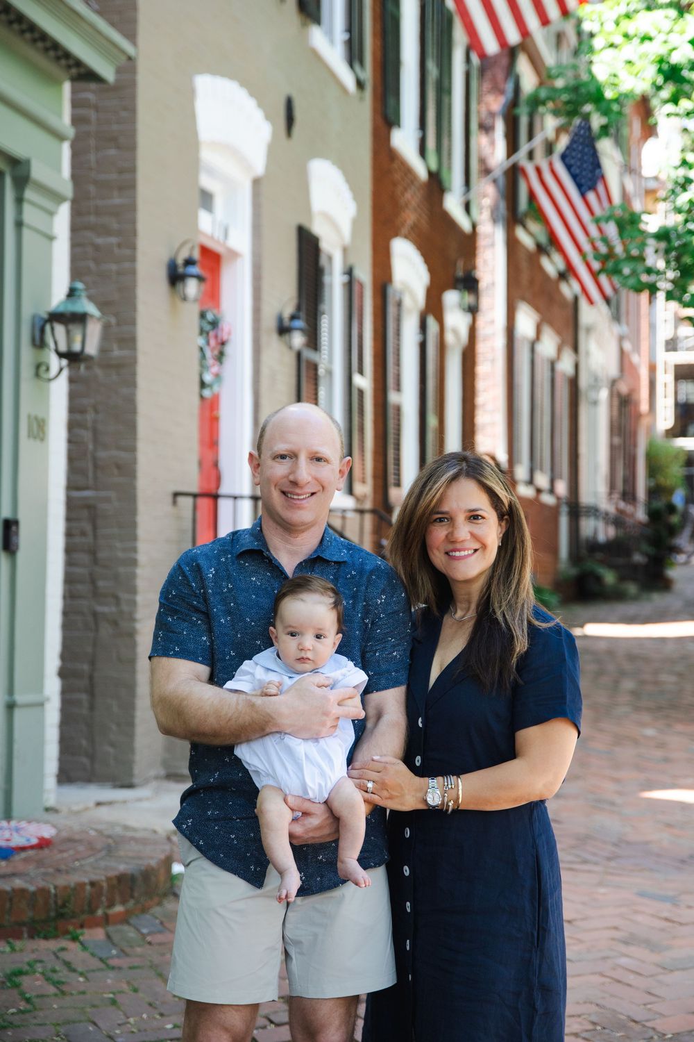 Happy couple in matching navy outfits holding their newborn baby on a historic brick sidewalk.