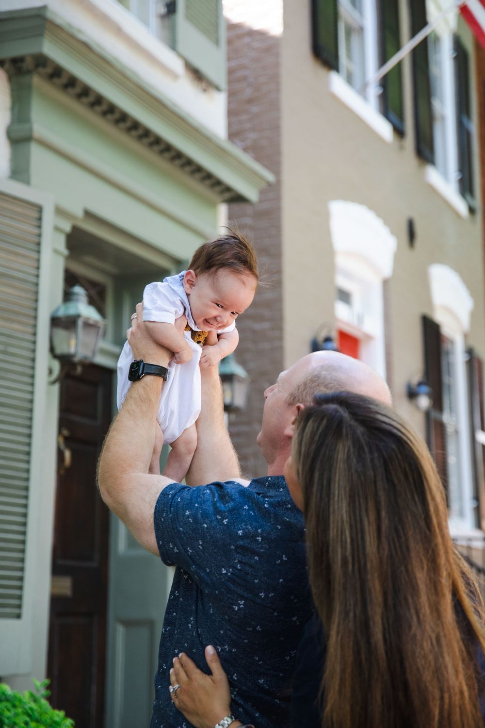 Parents joyfully lifting their smiling baby in the air outside a colorful townhouse.