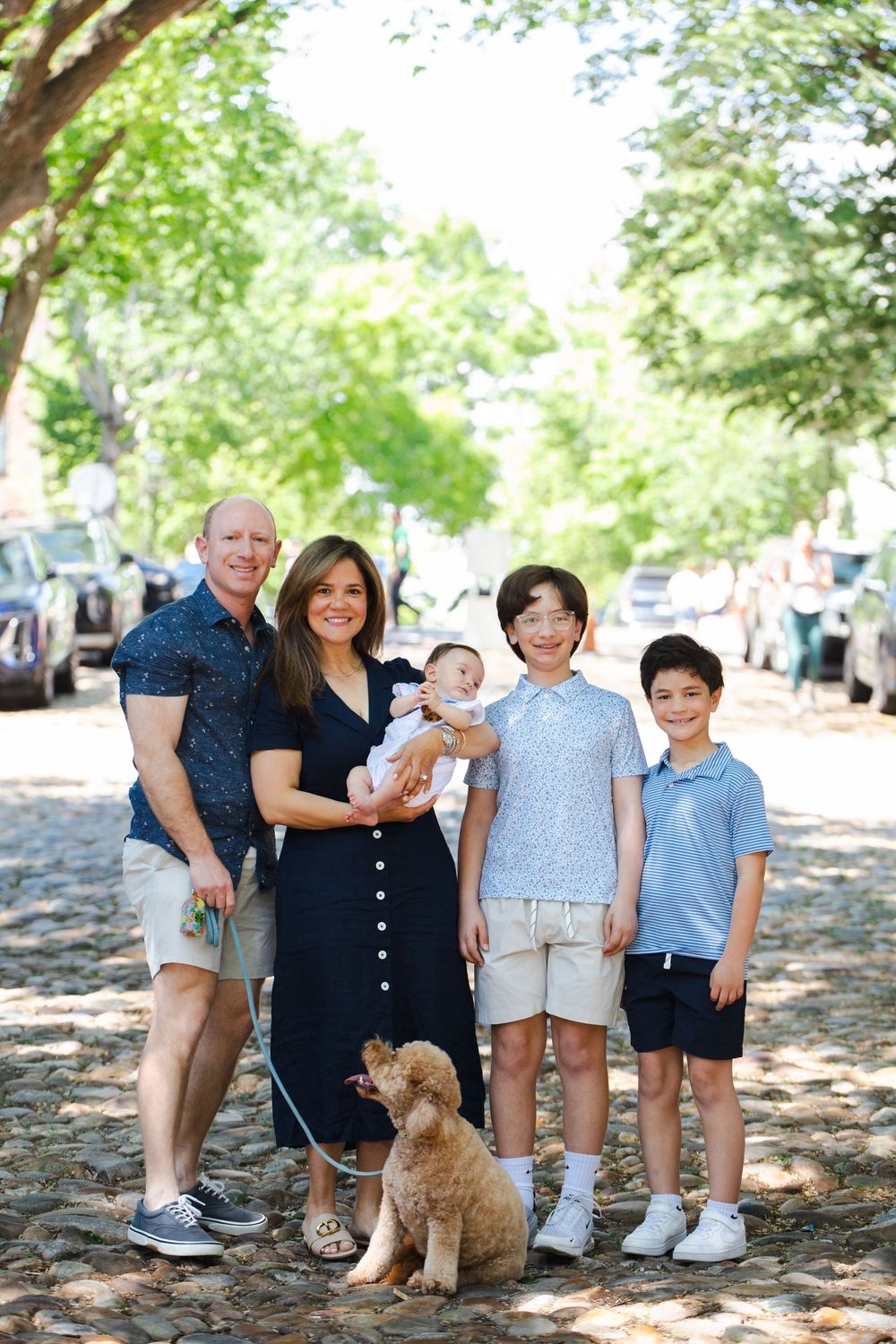 Family portrait with parents, three kids, and a golden doodle on a tree-lined cobblestone street.