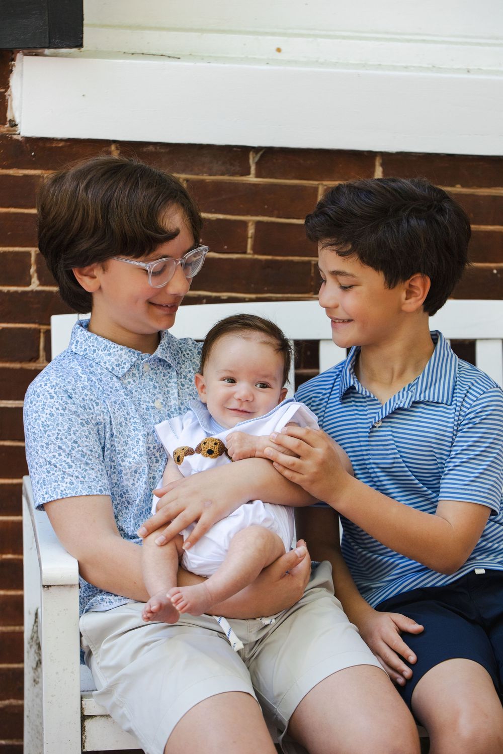Two boys sitting on a white bench holding a smiling baby on a brick porch.