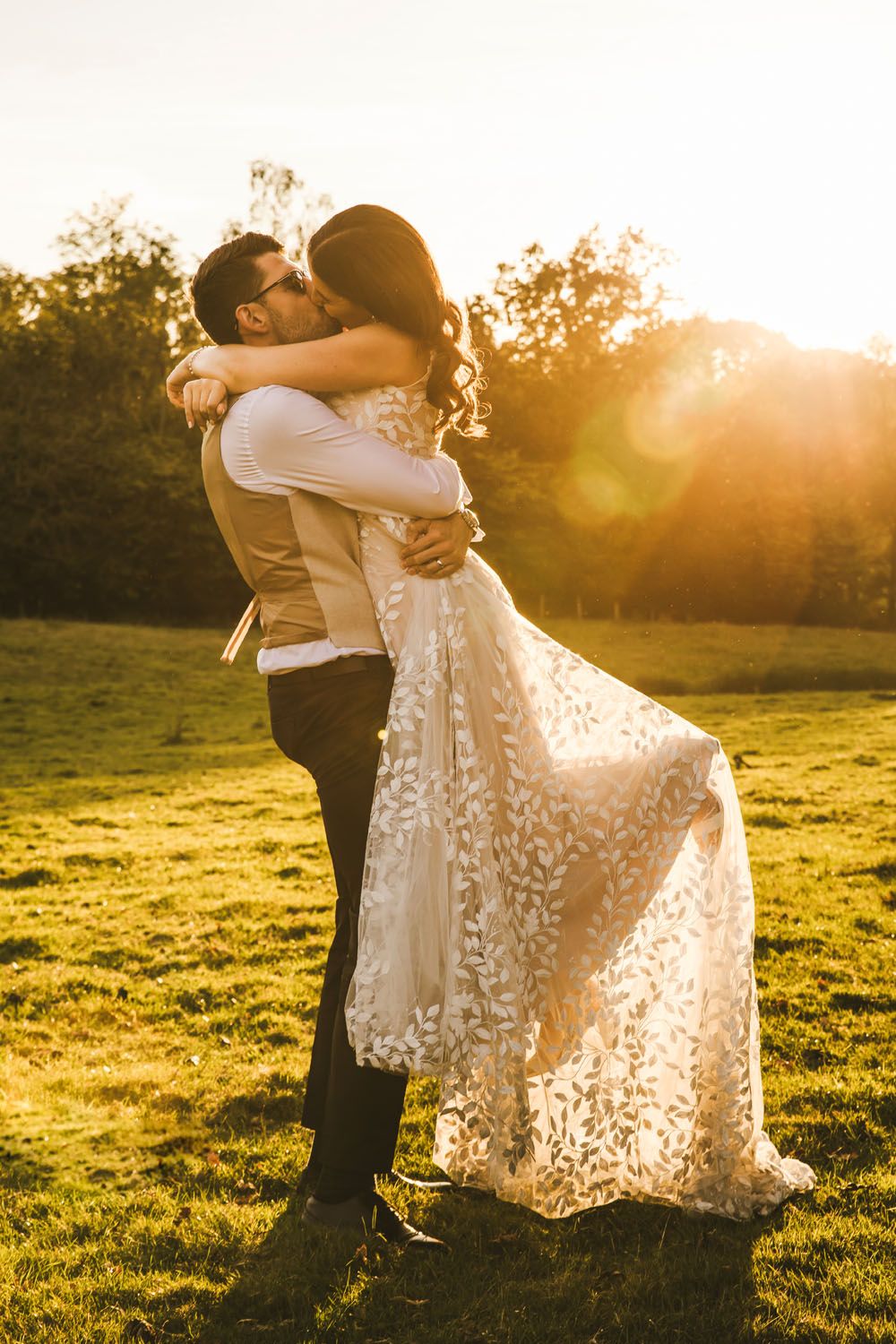 Bride and groom posing outdoors in golden sunlight, she in lace gown, he in vest and sunglasses.