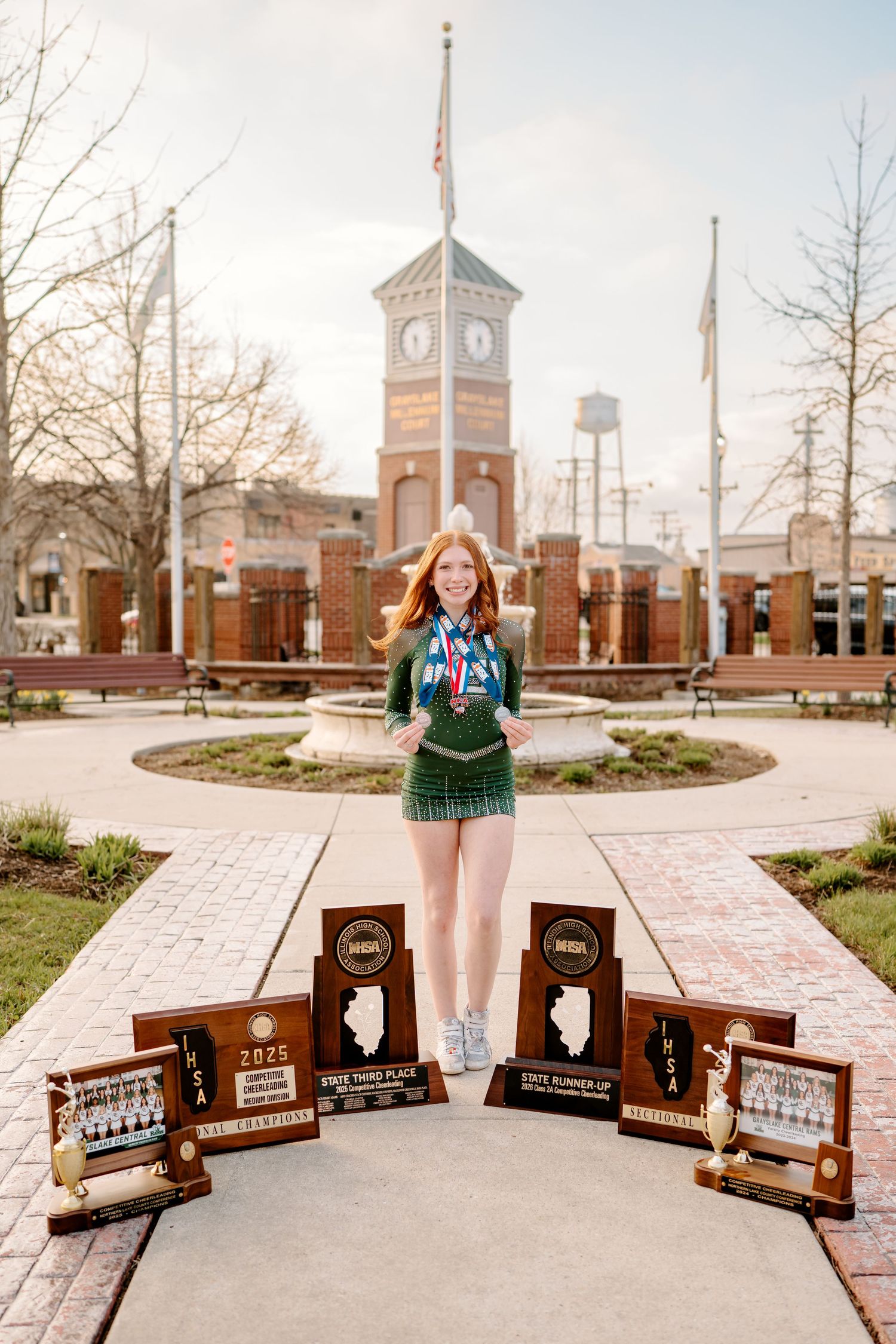 Red-haired girl in green outfit poses with state championship trophies and medals in front of brick clock tower.