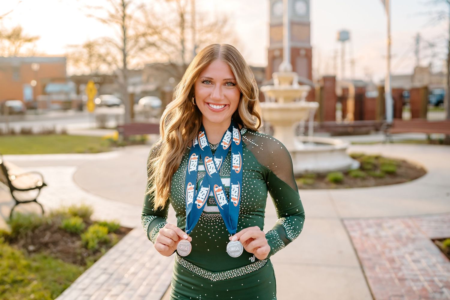 Woman in green sequin dress wearing beaded necklace smiling outdoors near a clock tower.