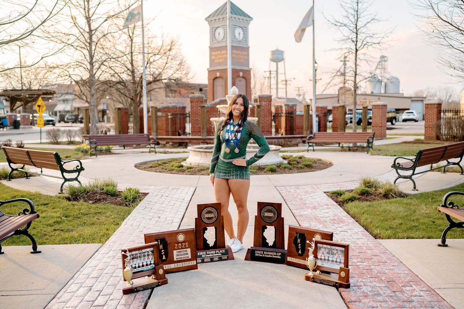 Young woman in green sweater poses with award plaques in a brick plaza featuring a clock tower backdrop.