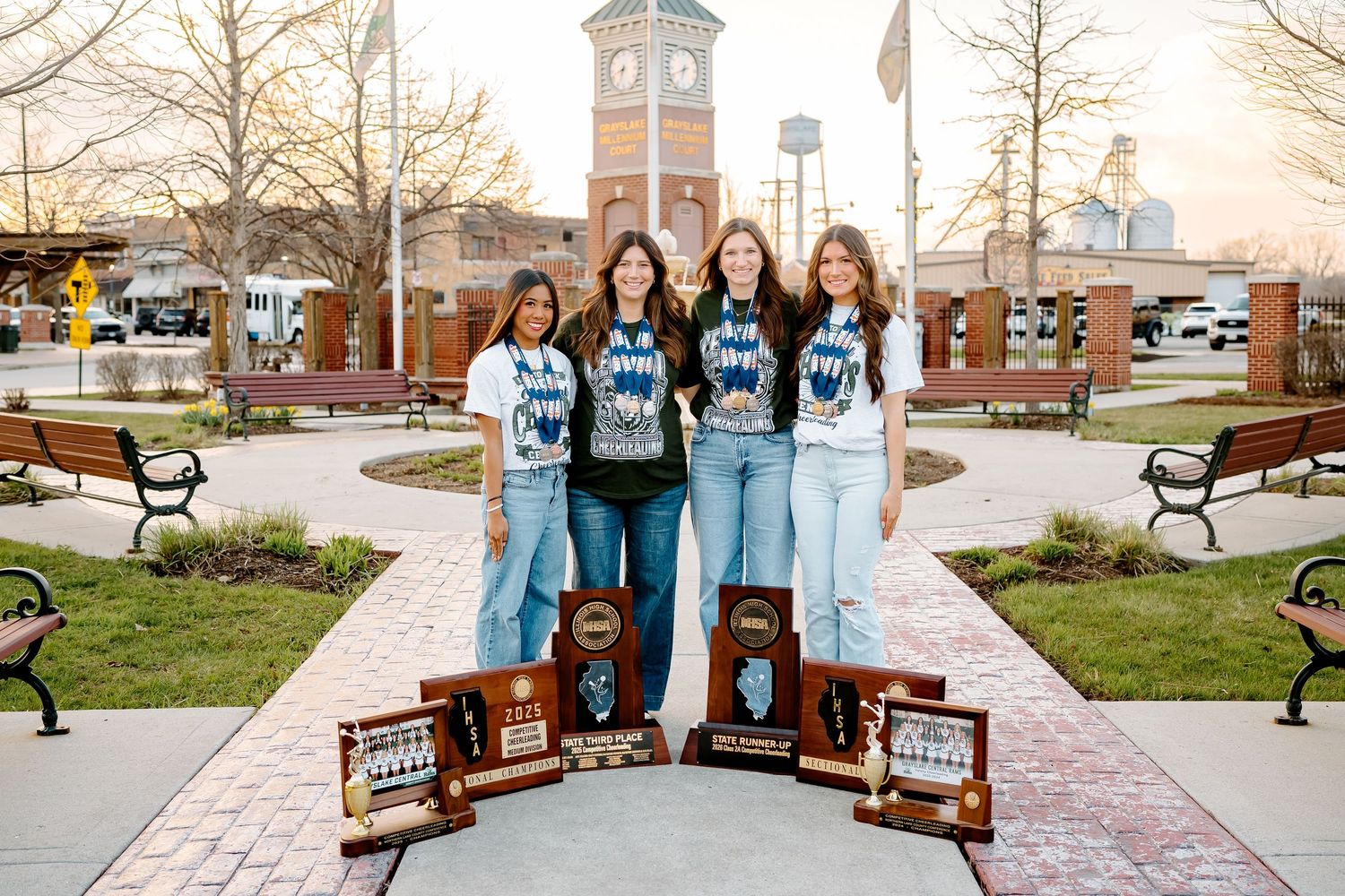 Four young women pose with championship trophies outdoors near a clock tower, wearing matching blue team shirts.
