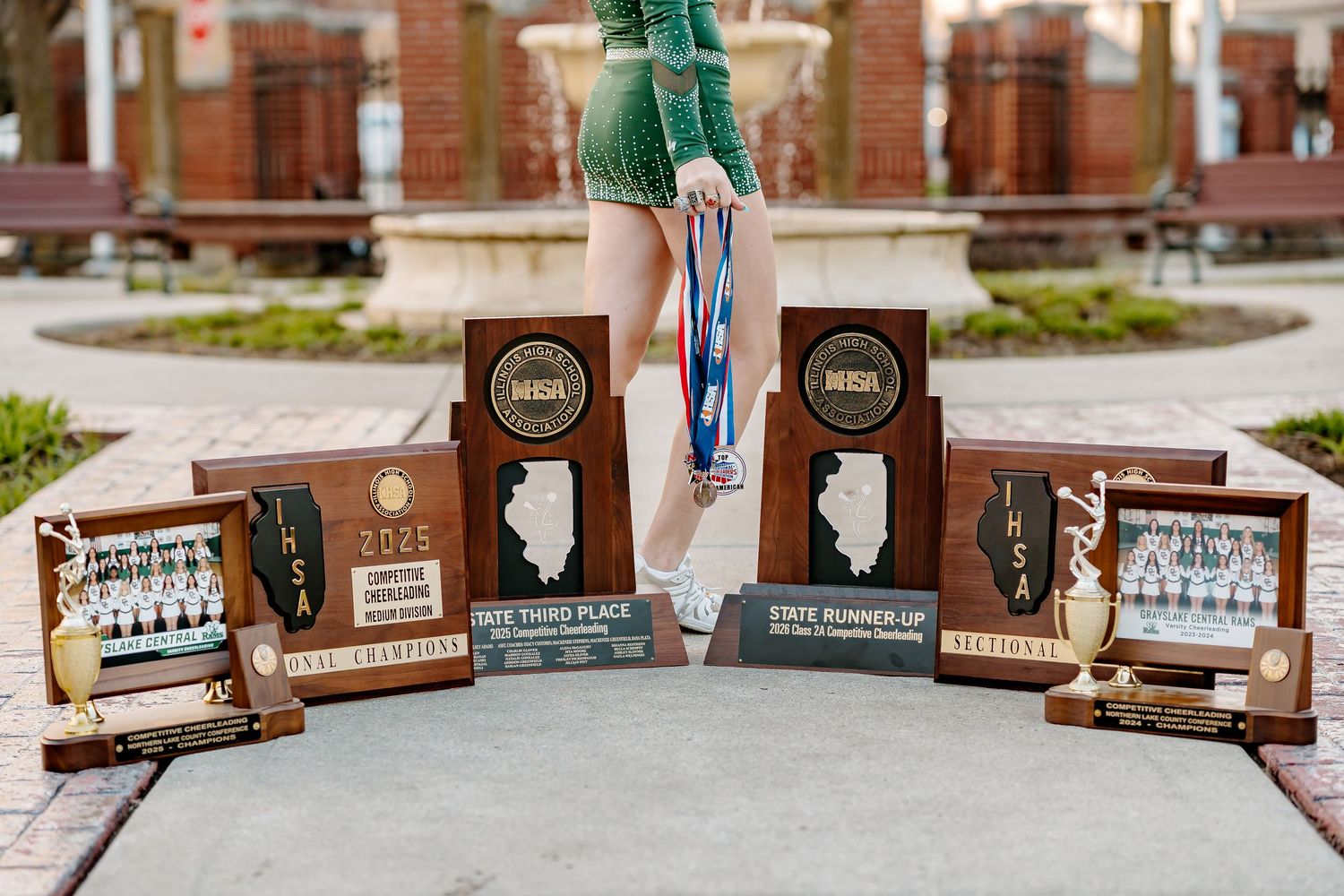 Gymnast displays IHSA state trophies including third place and state runner-up awards with medals on outdoor brick steps.