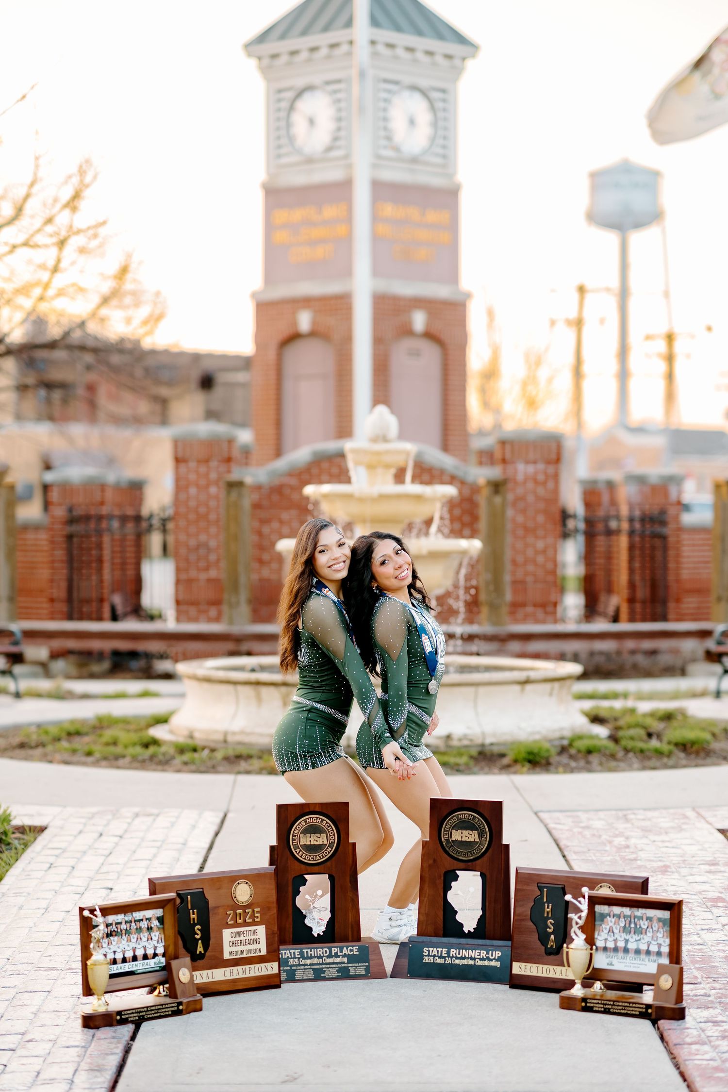 Two girls in green outfits pose back-to-back with state championship trophies displayed in front of brick clock tower.