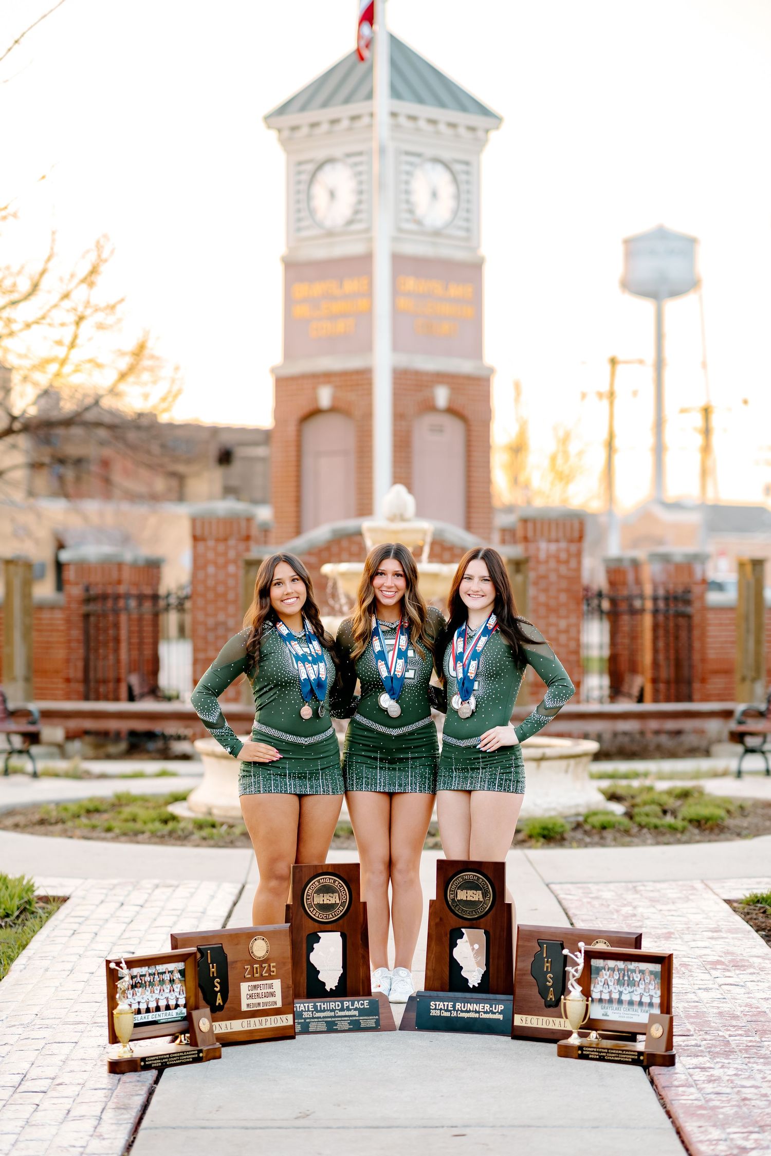 Three young women in green uniforms display championship trophies and medals near a brick clock tower.