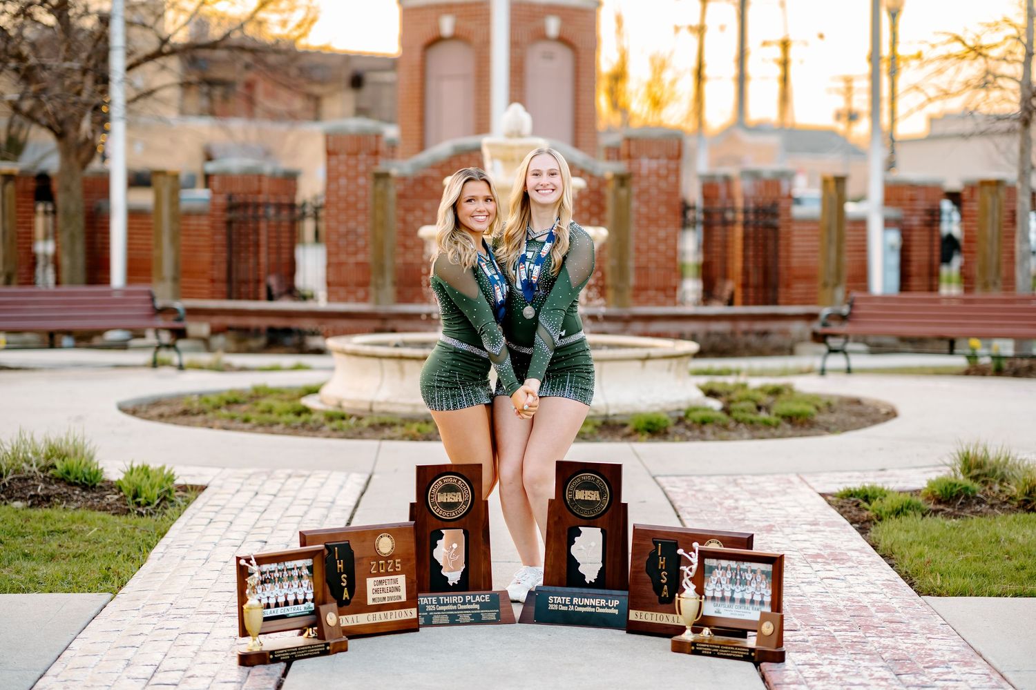 Two young women in green jackets pose with multiple trophies and awards on a brick plaza near a fountain.