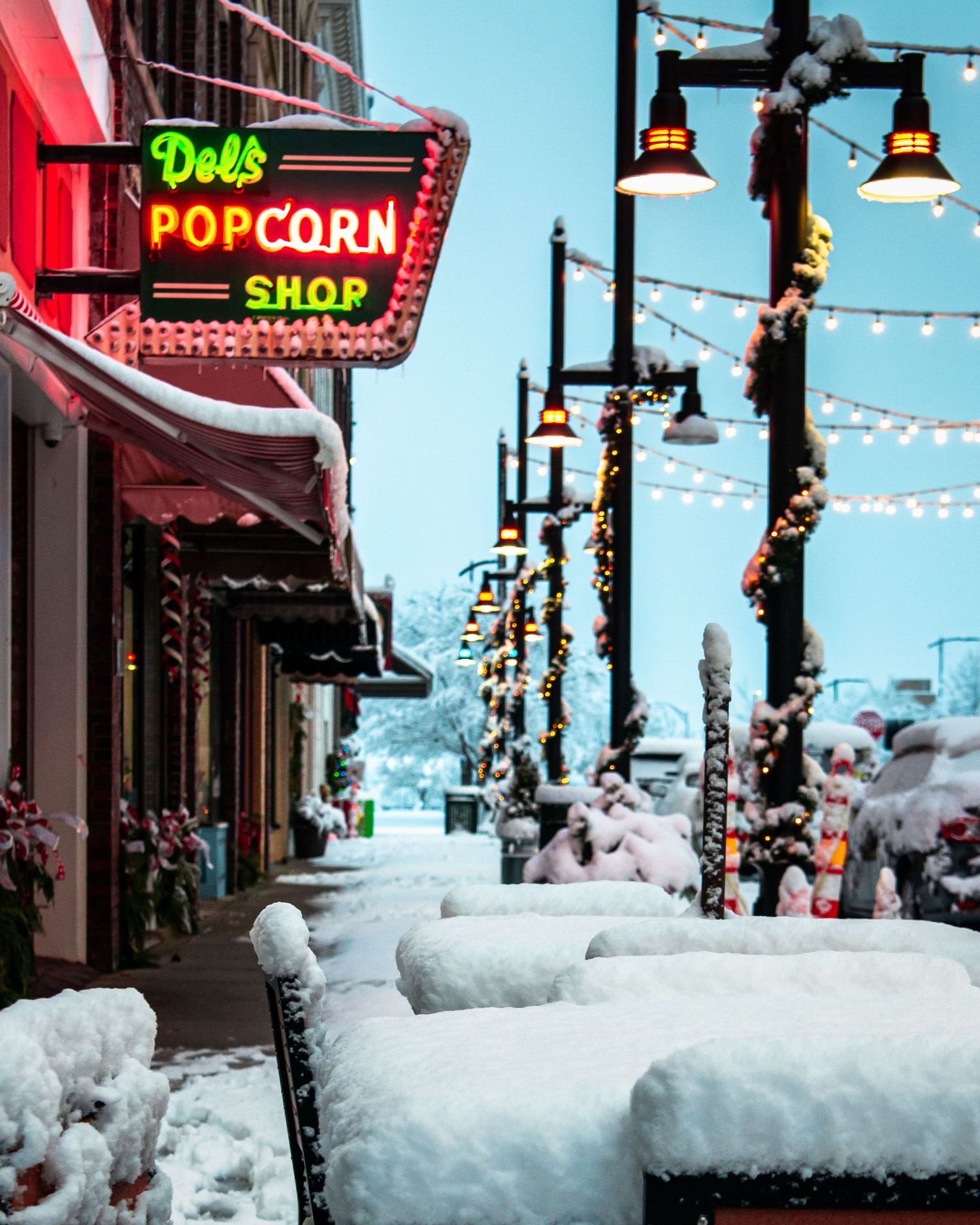 Snow-covered street with Del's Popcorn Shop neon sign, string lights, and festive winter decorations lining a charming small town.