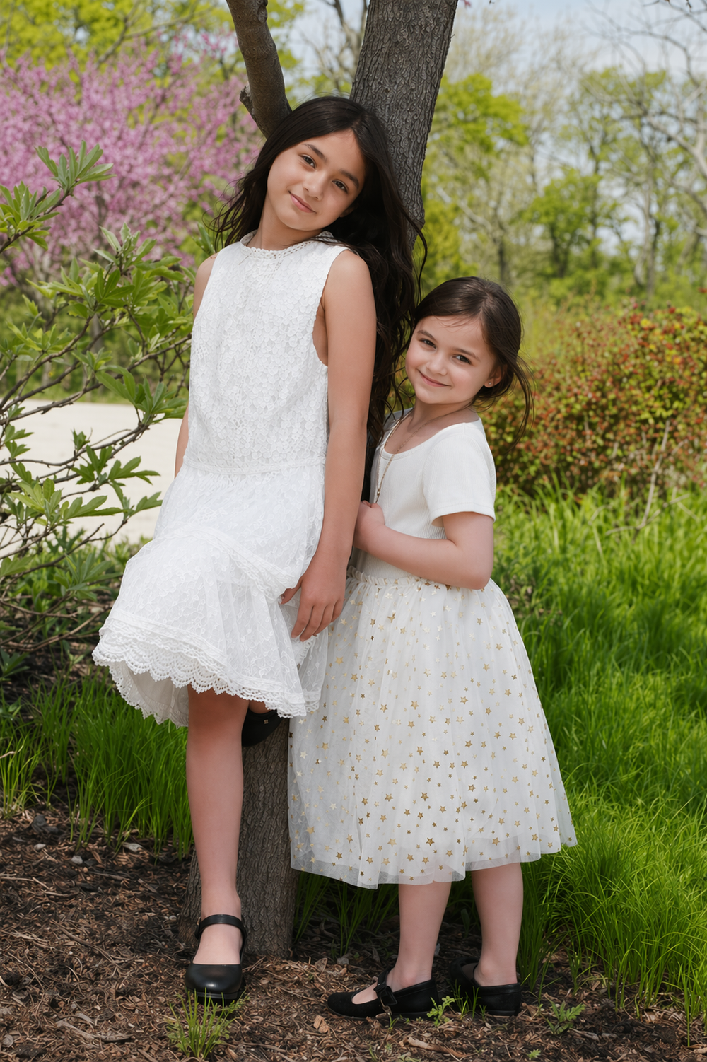Two young girls in white dresses pose together by a tree in a lush green garden with blooming flowers.