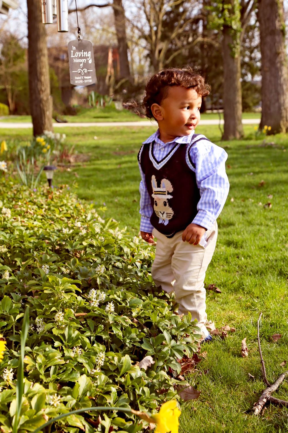 Toddler boy in Easter vest and plaid shirt standing among spring flowers in a lush green park.