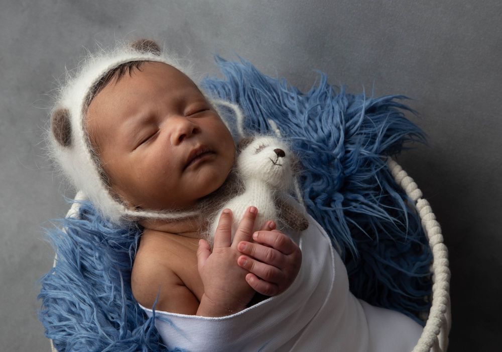 Sleeping newborn baby in white wrap with bear bonnet, holding a teddy bear in a blue fur basket.