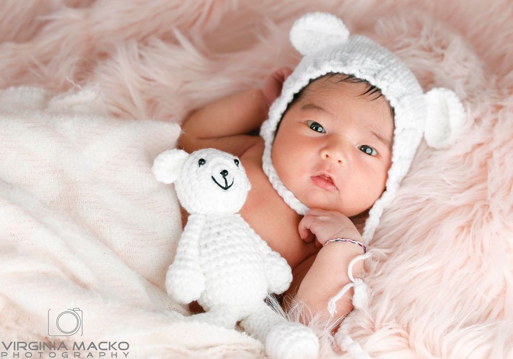 Newborn baby in white bear bonnet hat lying on pink fur blanket with matching crochet teddy bear toy.