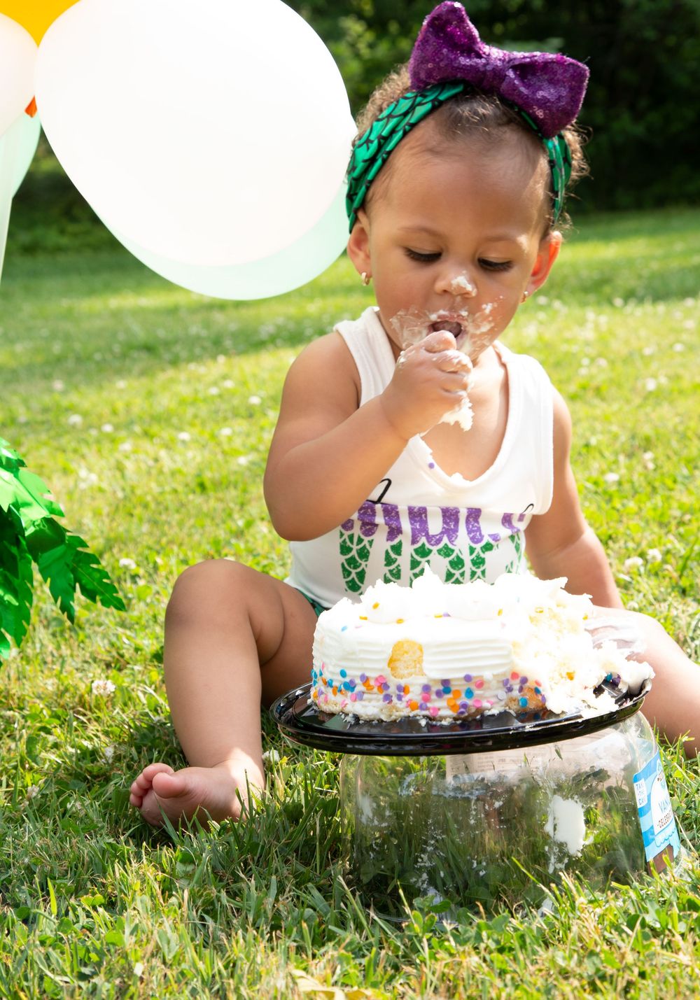 Toddler girl in white onesie and purple bow headband smashing birthday cake outdoors on green grass with balloons.