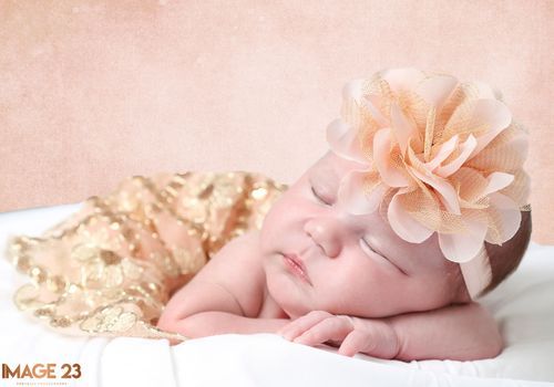 Sleeping newborn baby wearing a peach floral headband, wrapped in a gold sequin blanket on a white surface.