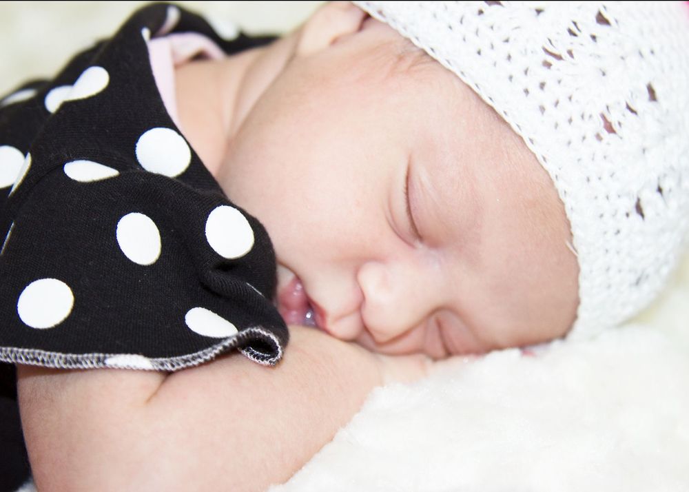 Sleeping newborn baby wearing a white knit hat and black polka dot outfit, resting peacefully on a soft white blanket.
