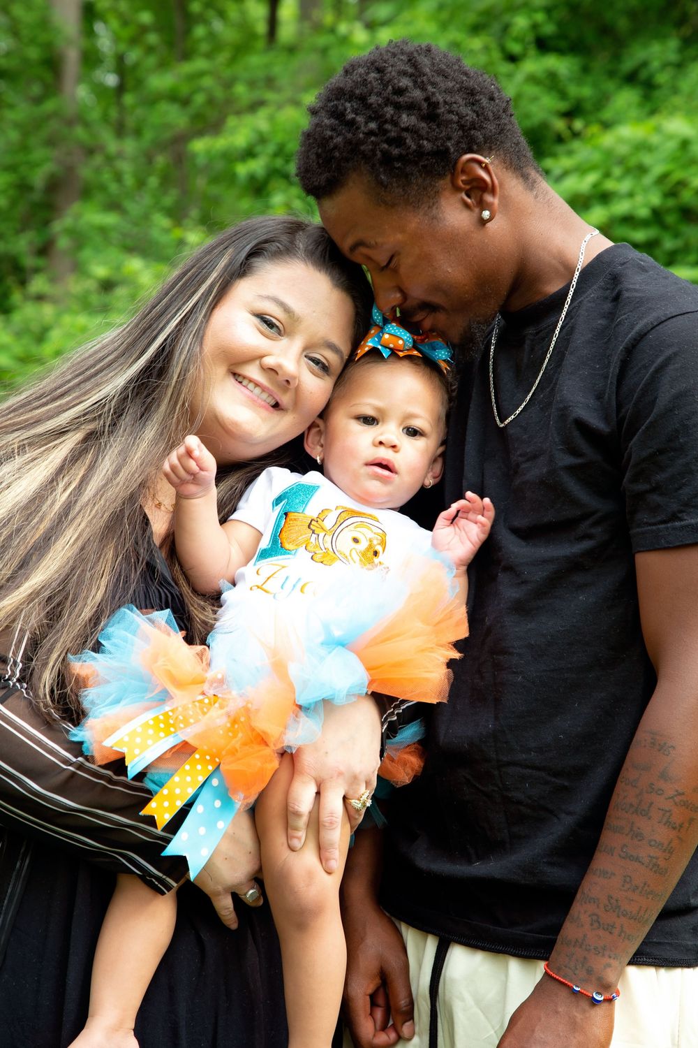 A smiling couple holds their baby girl in an orange and teal tutu outdoors with green trees behind them.