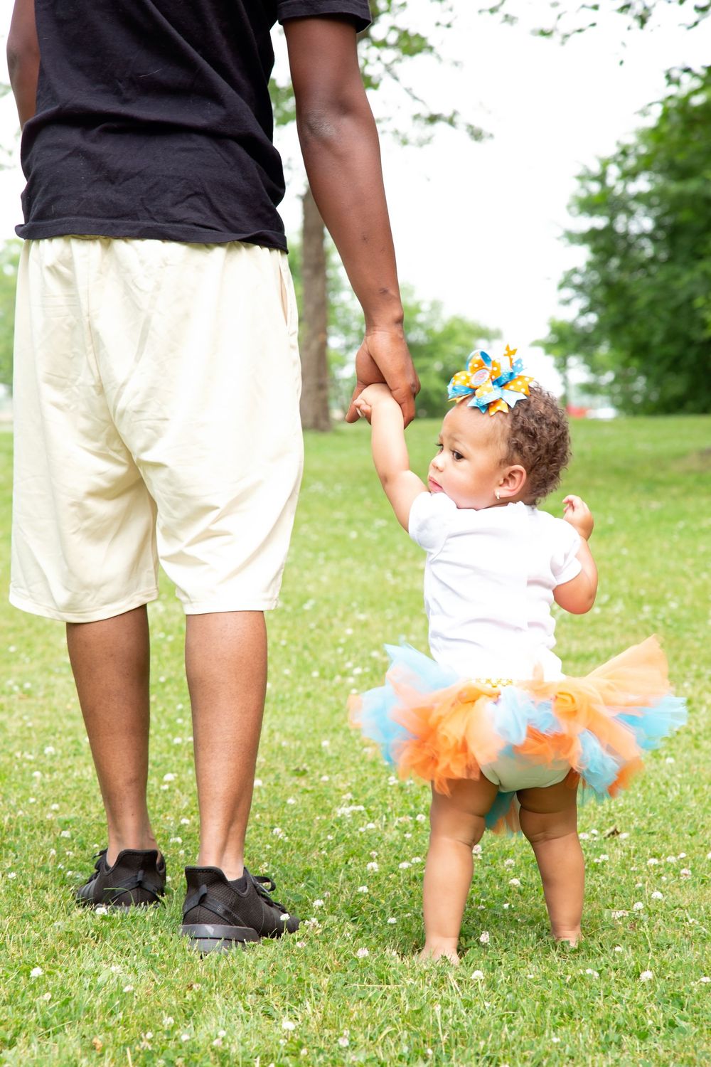 A toddler in a colorful tutu holds an adult's hand while learning to walk on green grass outdoors.