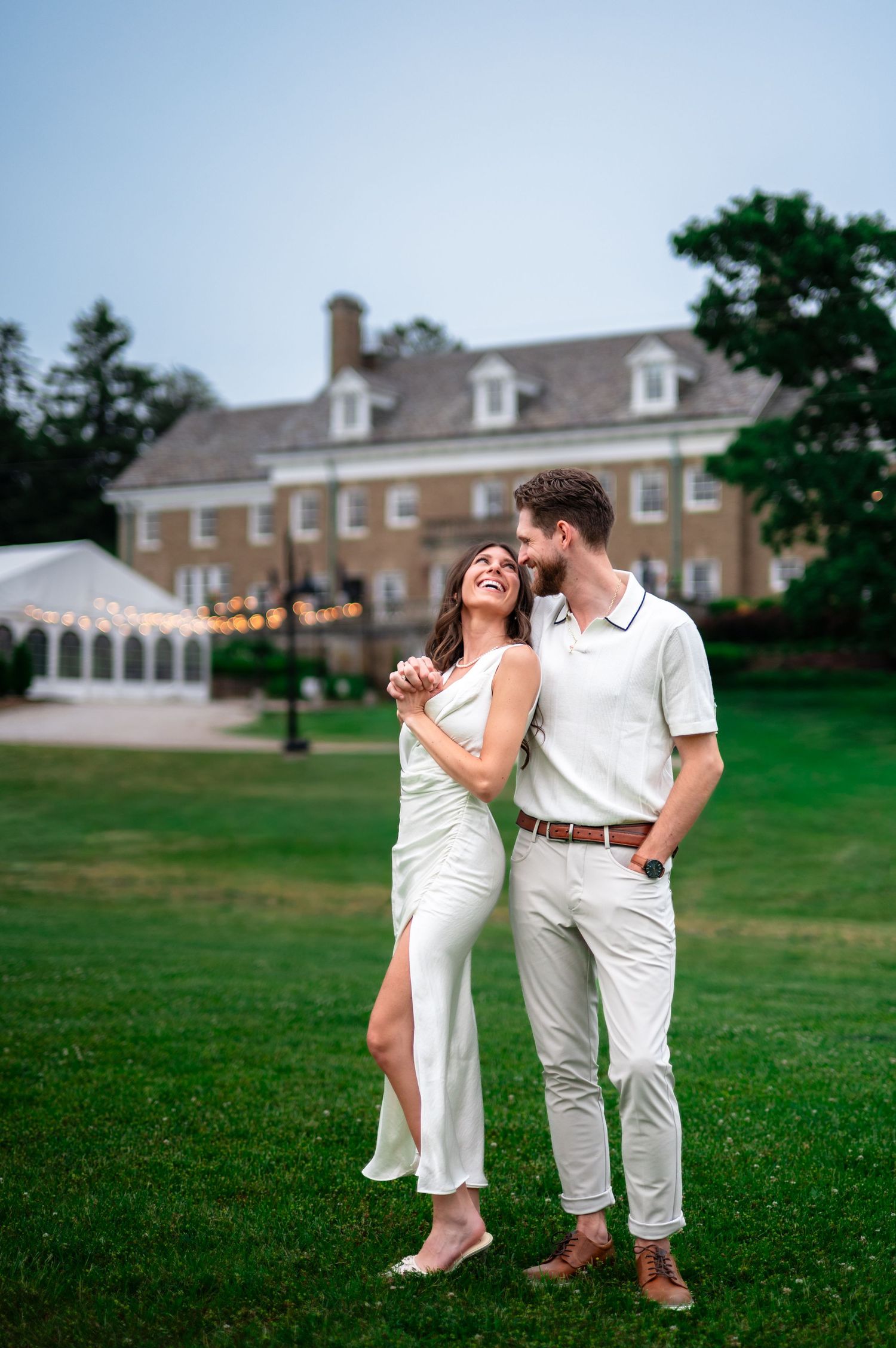 Couple in white outfits embracing on green lawn before elegant brick manor at dusk.
