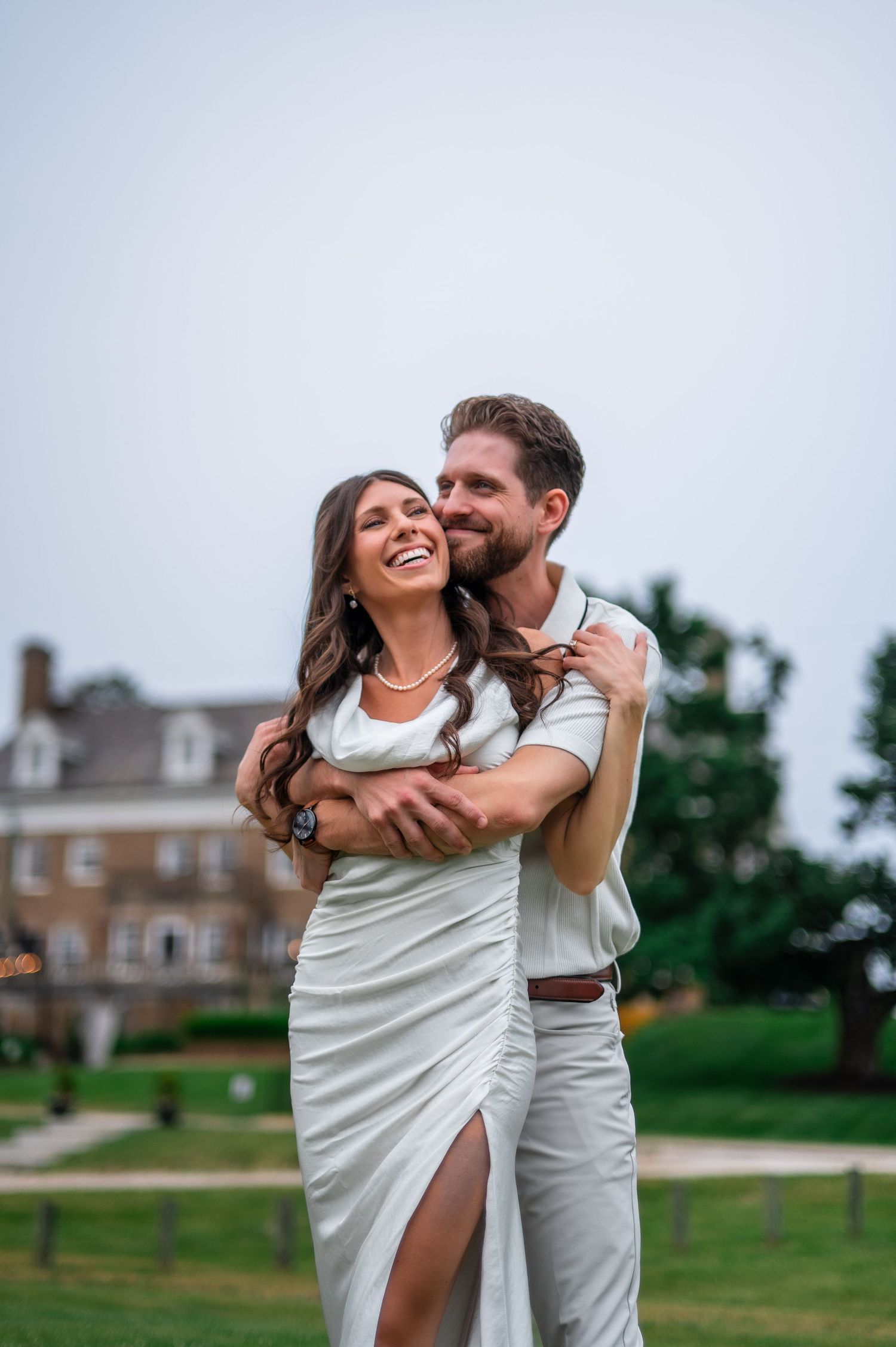 Smiling couple in white outfits embracing outdoors near a brick building on a green lawn.