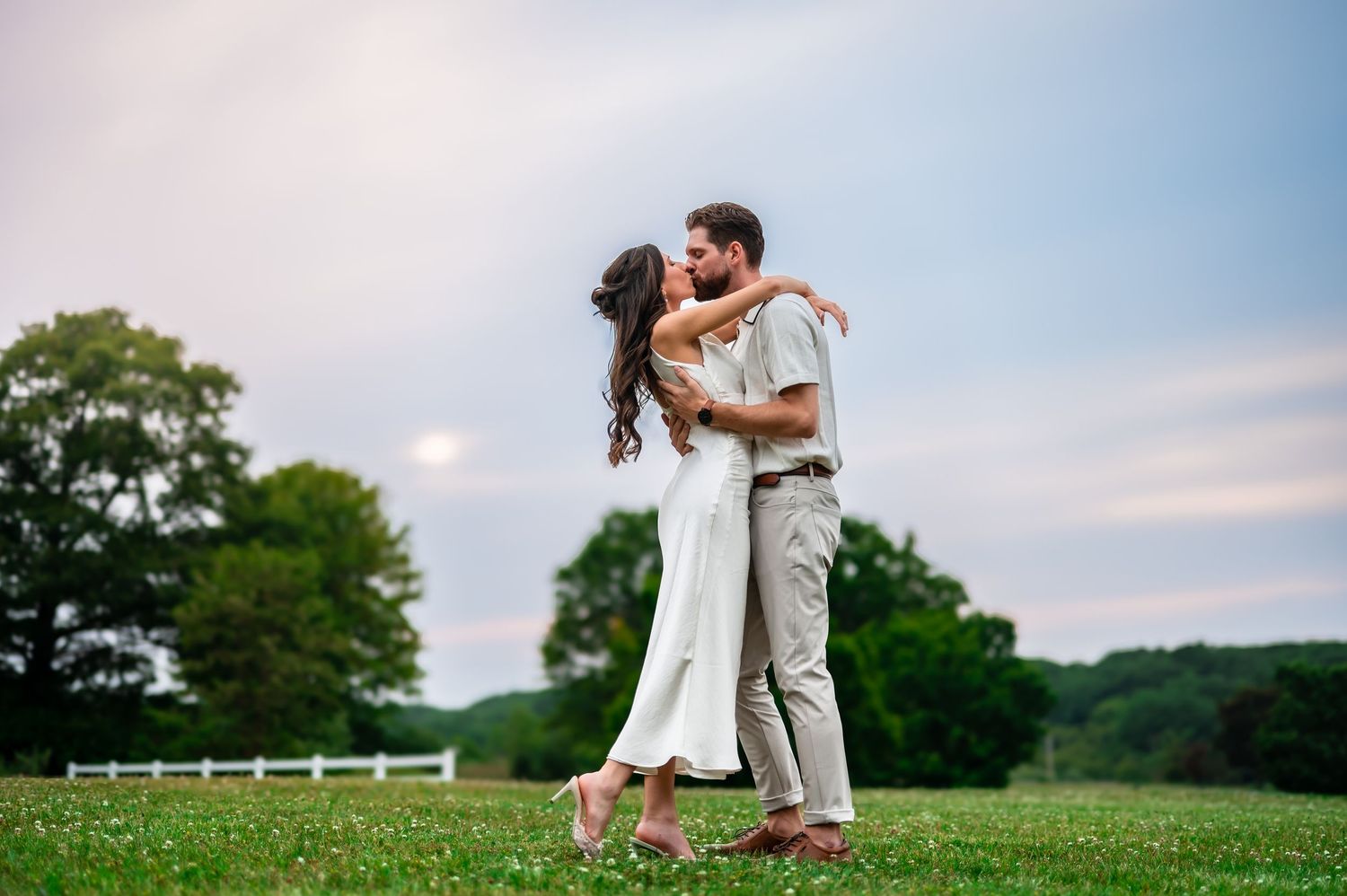 Couple sharing a romantic kiss in a lush green meadow at dusk, woman in white dress, man in casual attire.