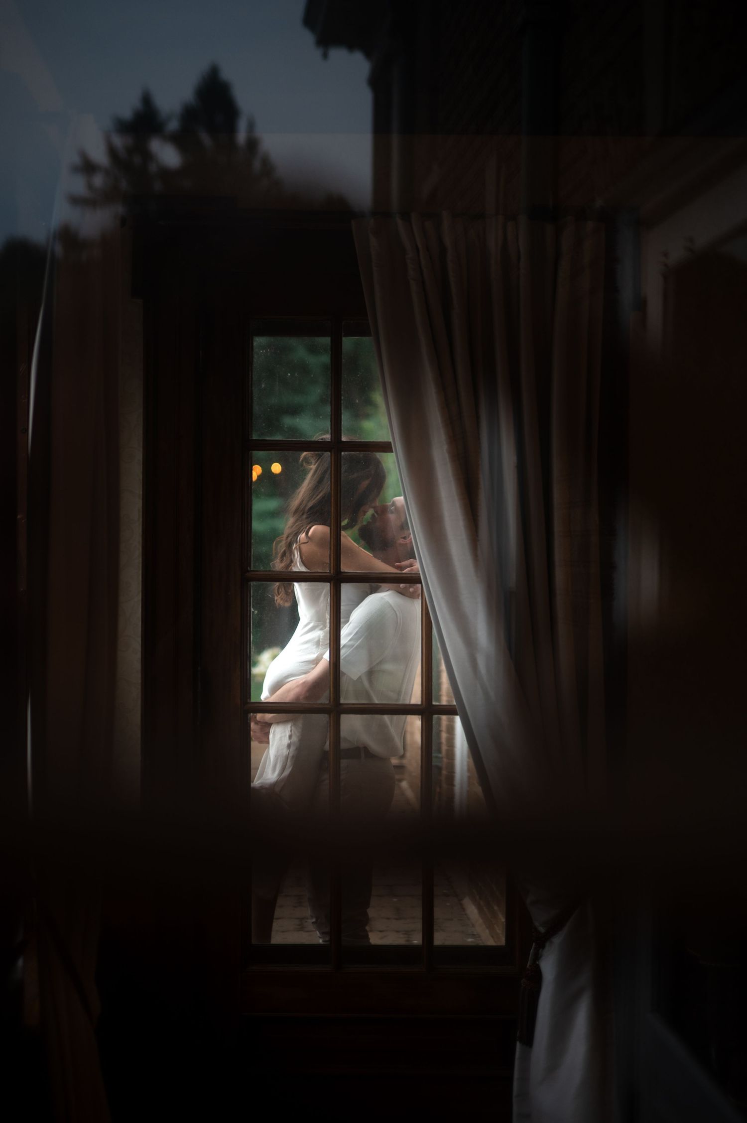 Romantic couple silhouetted kissing through window with sheer curtains at night.