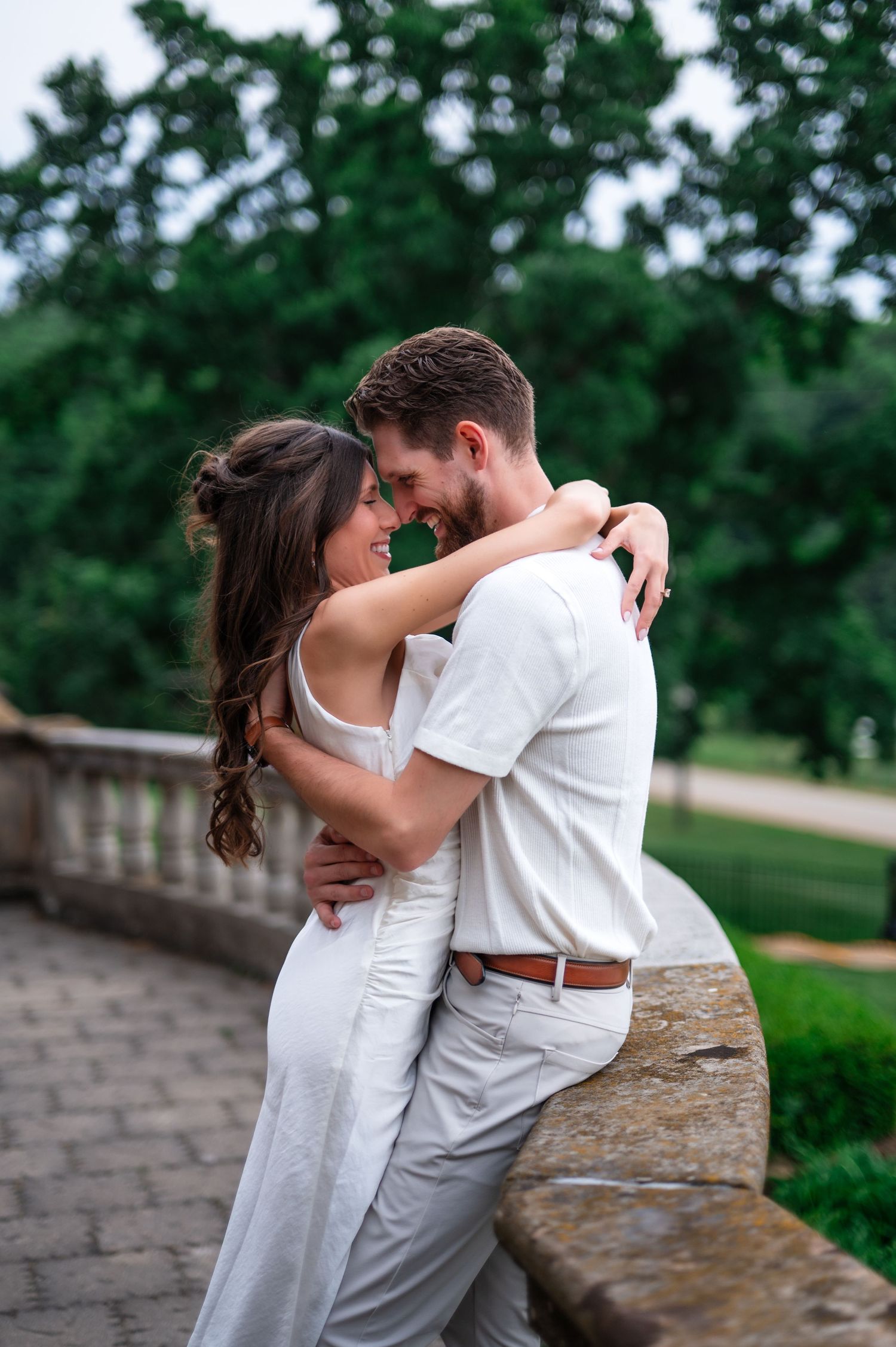 Couple in white embracing closely on a stone balustrade surrounded by lush green trees.