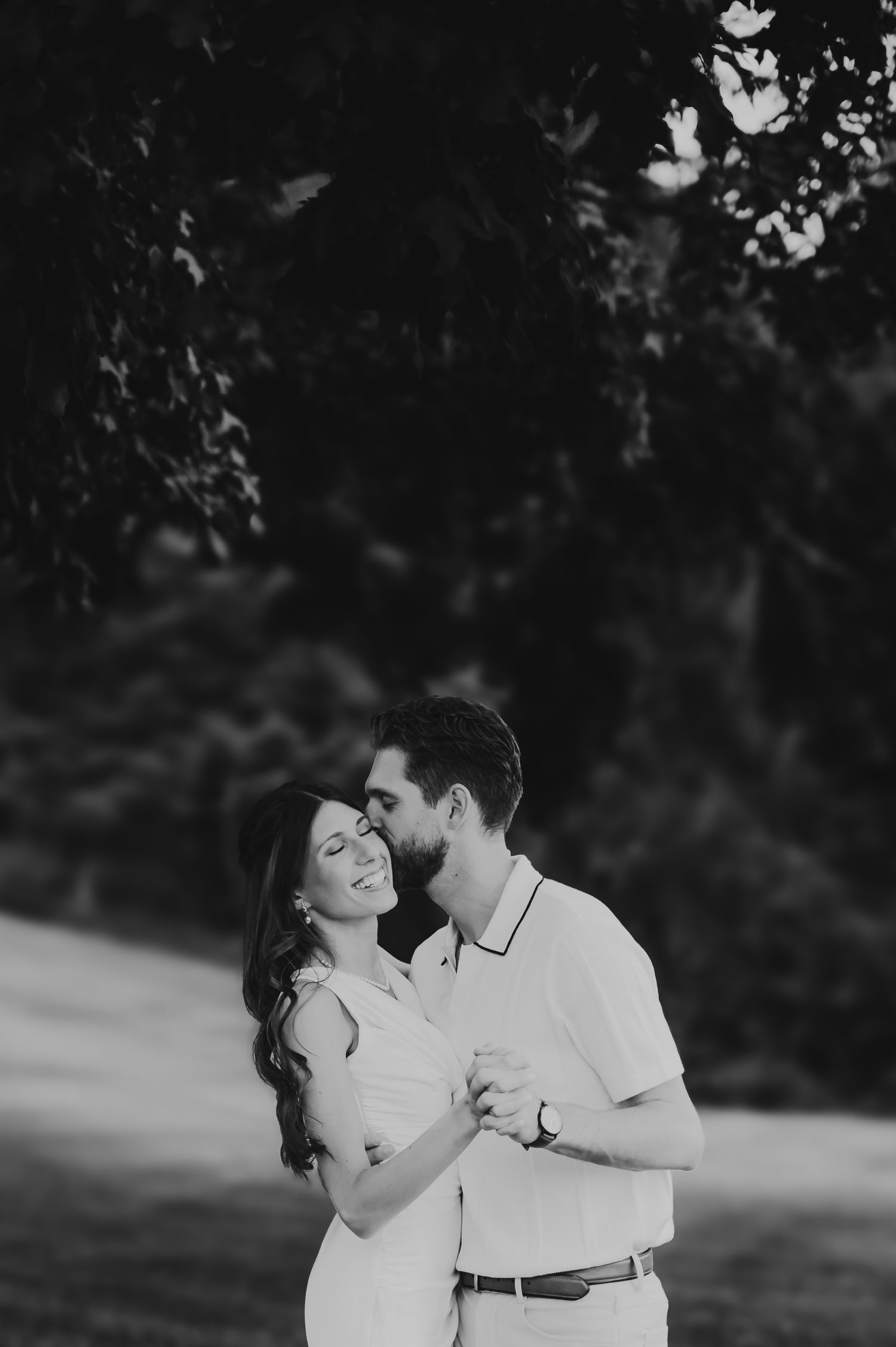Black and white photo of a romantic couple embracing and dancing outdoors among lush trees.