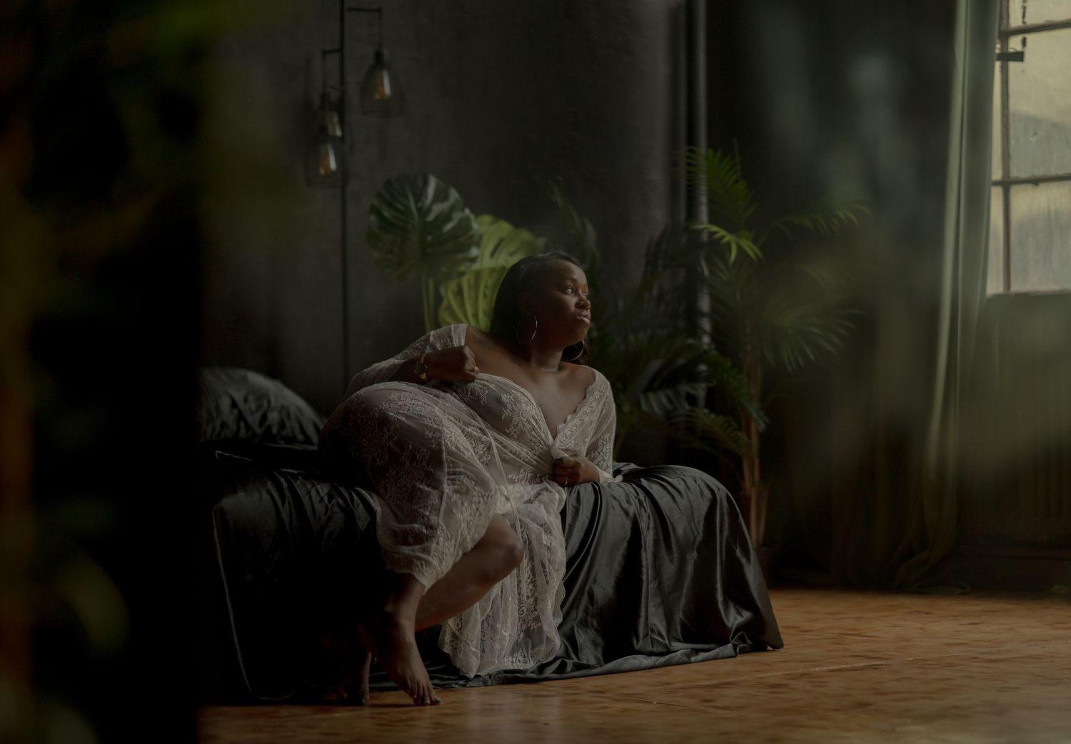 A woman in a white lace dress sits pensively on a dark sofa in a moody, plant-filled room with soft natural light.