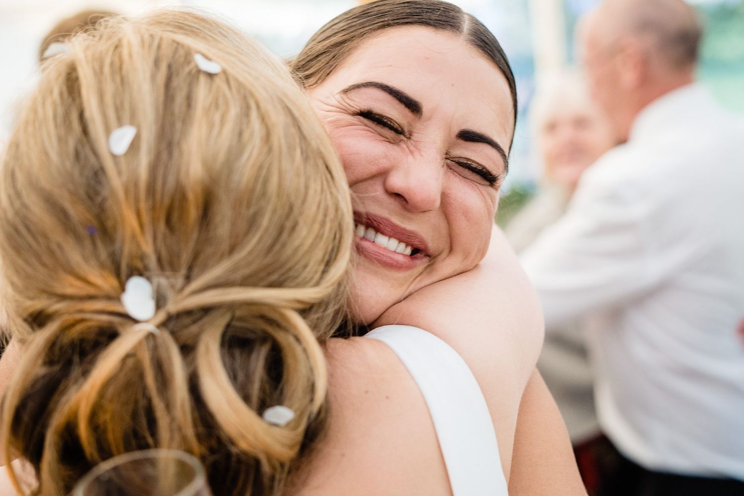 Two women sharing a warm embrace at a wedding, with the bride smiling joyfully as guests celebrate in the background at Oceanstone Resort