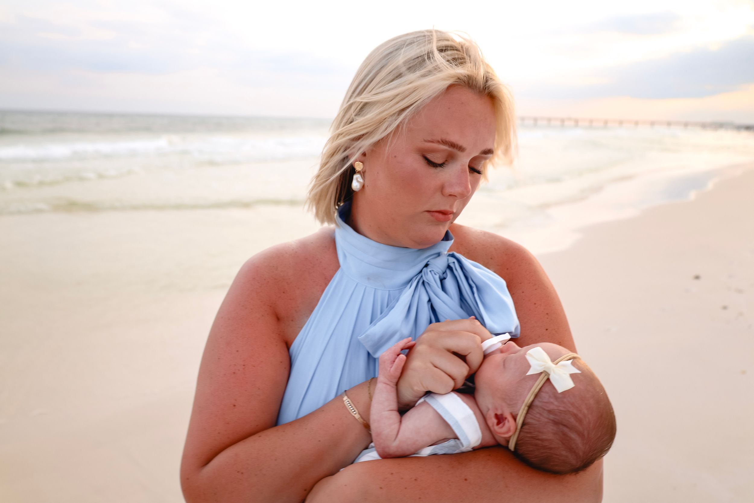 A mother in a blue dress holds her newborn baby on a sandy beach at sunset.