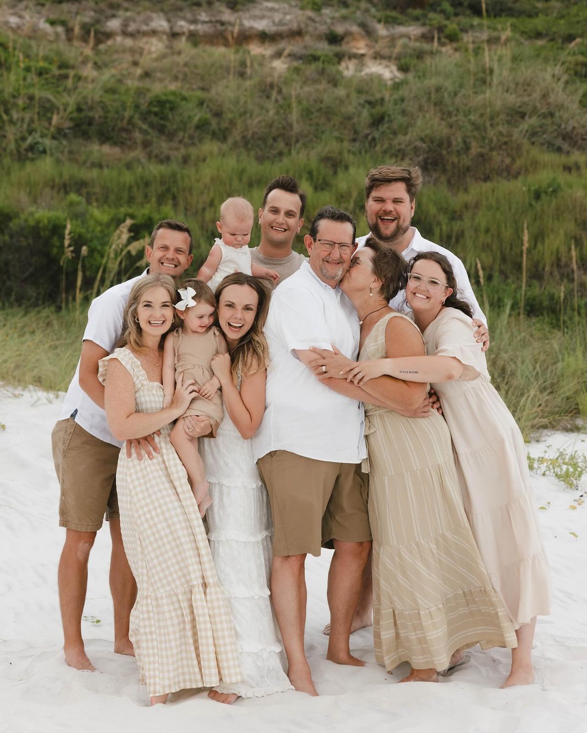 A large happy family poses together on a white sandy beach surrounded by lush green dunes.