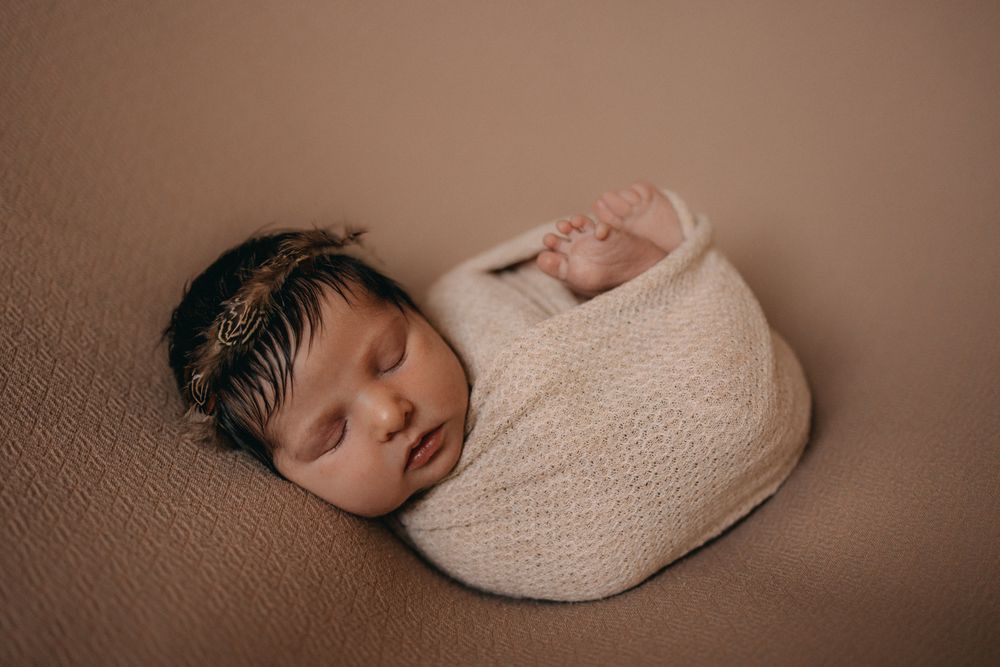 Newborn baby swaddled in white knit wrap sleeping peacefully on beige blanket with delicate gold headband.