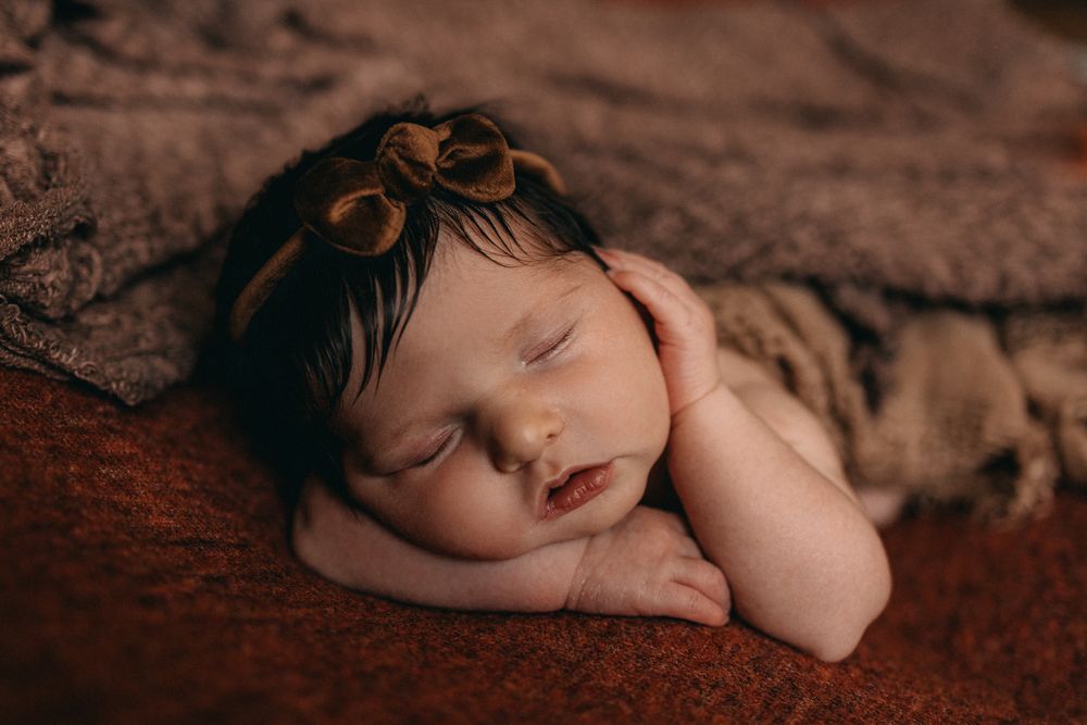 Peaceful newborn baby girl sleeping with dark hair and bow headband on a soft rustic brown blanket.
