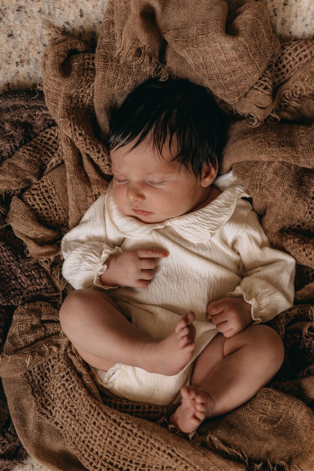 Peaceful newborn baby with dark hair sleeping in a cream onesie on rustic burlap fabric.
