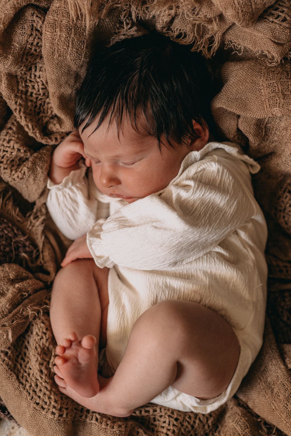 Newborn baby sleeping peacefully in white romper on rustic burlap fabric, curled in natural fetal position.