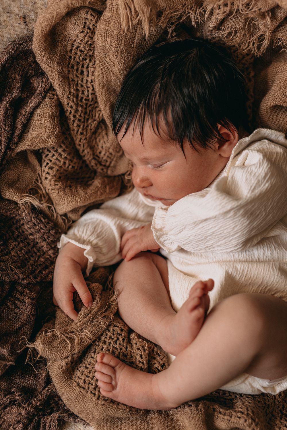 Peaceful newborn baby sleeping on brown textured blanket, wearing white romper with dark hair.