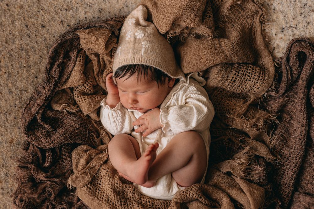 Sleeping newborn baby wearing a knit beige gnome hat, wrapped in brown textured blankets in a cozy nest.