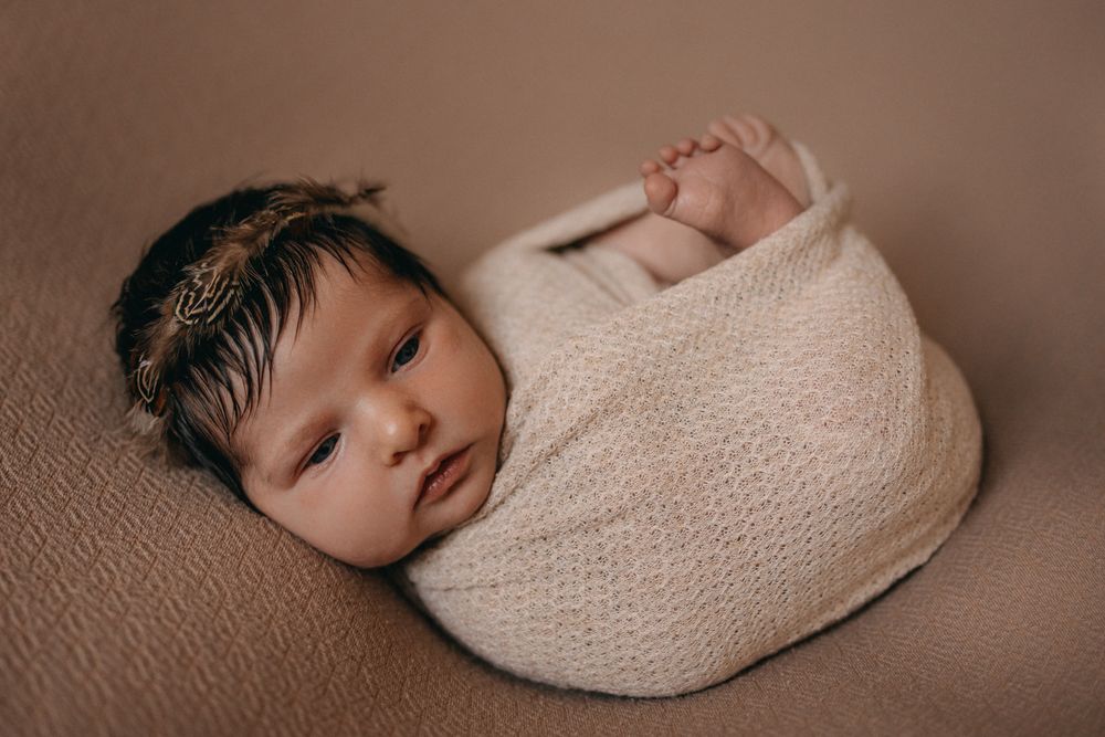 Newborn baby swaddled in cream knit wrap with floral headband, peacefully resting on brown backdrop.