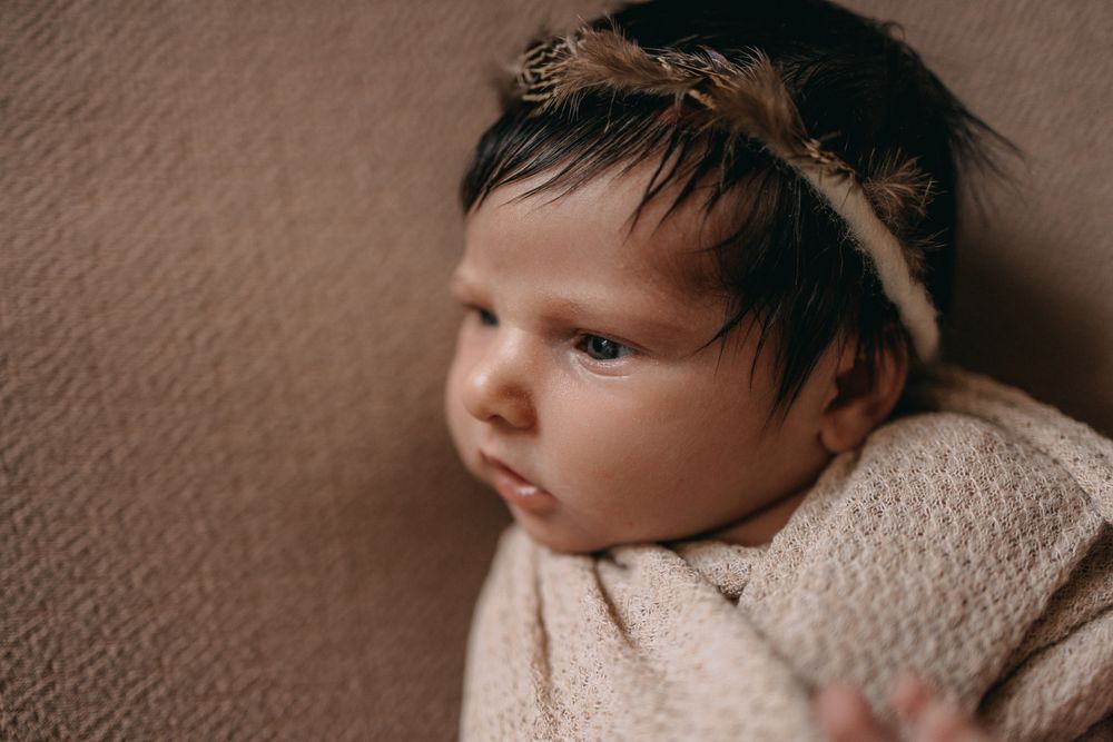 Newborn baby wrapped in a soft beige blanket wearing a delicate feather headband, gazing peacefully to the side.