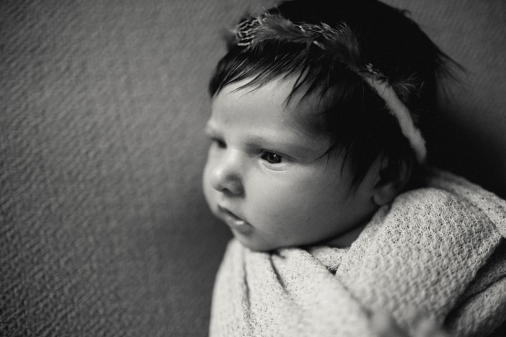 Black and white newborn portrait of a dark-haired baby wrapped in soft blanket wearing a delicate headband.