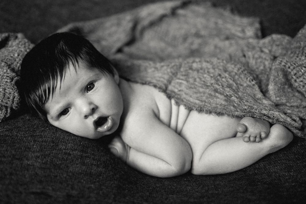 Black and white newborn baby photo, infant lying on soft blanket with dark hair and wide eyes.
