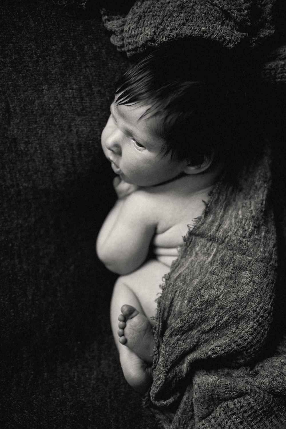 Black and white photo of a sleeping newborn baby wrapped in a knit blanket, curled up peacefully.