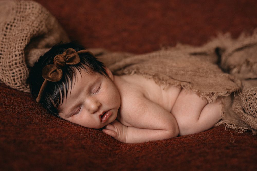Peaceful newborn baby girl sleeping wrapped in burlap with a brown bow headband on a rustic red backdrop.